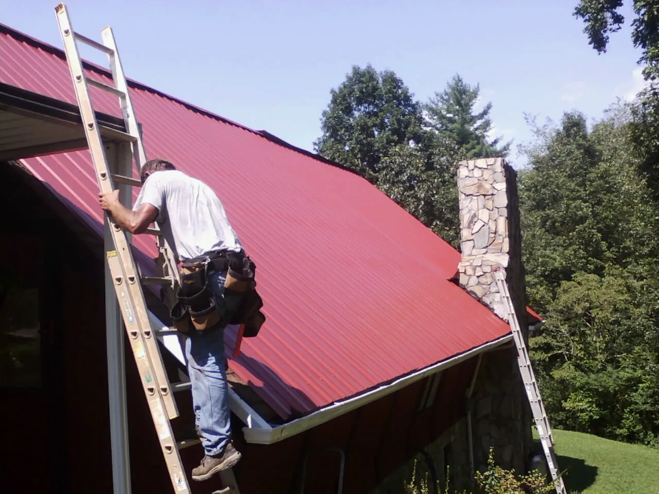 Roofer on a ladder beside a red metal roof and stone chimney — Soffit & Fascia Repair by Roofing Brothers in Towaco